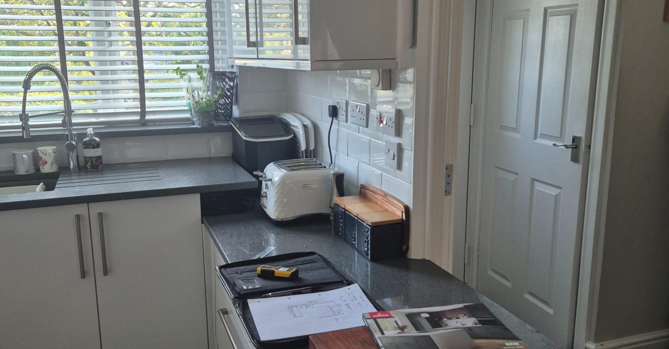 Existing white kitchen in Stamford with gloss cabinets, dark worktops, a window sink and design samples laid across the work surface.