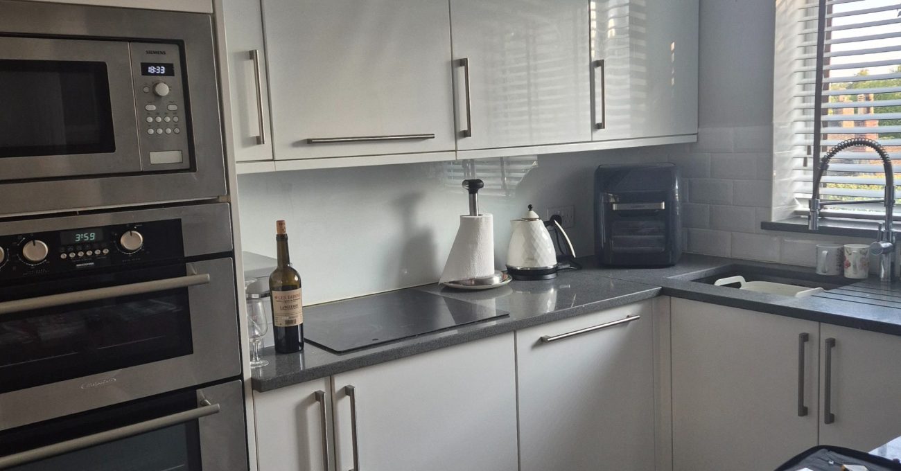Original kitchen cooking area with white gloss units, stacked ovens, dark worktops and wall cabinets beside the window.