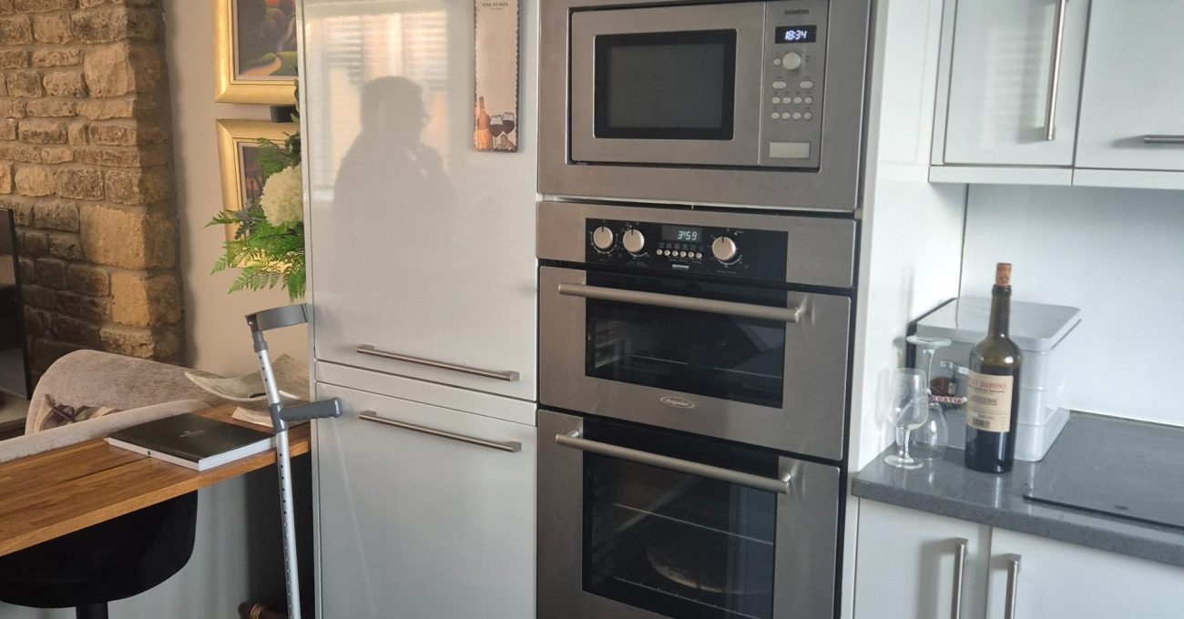 Tall white kitchen cabinets and stacked appliances beside an exposed stone feature wall before the renovation began.